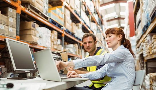 A man and woman sit at a desk in racking aisle with a laptop before them. The woman points at the screen.
